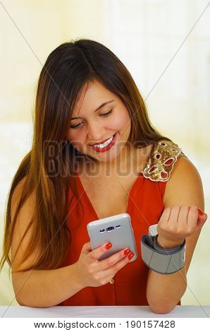 Beautiful young patient using an automatic tensiometer in her arm and texting in her cellphone, in a doctor consulting room background.