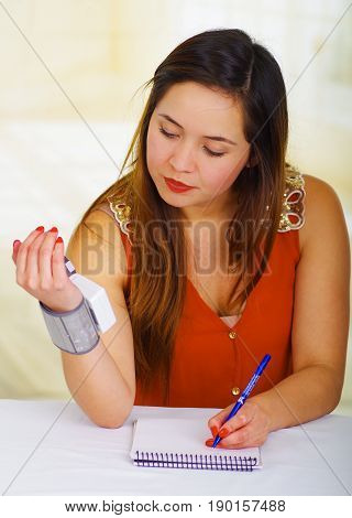 Beautiful young patient using an automatic tensiometer in her arm and taking notes of the results in his notebook, in a doctor consulting room background.