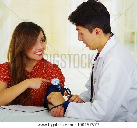 Handsome surprised young doctor with stethoscope around his neck taking the pulse to a young woman, in a doctor consulting room background.