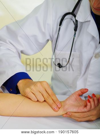 Handsome young doctor with stethoscope around his neck taking the pulse to a young woman, in a doctor consulting room background.