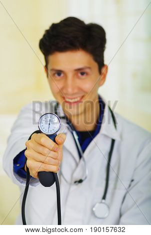 Handsome smiling young doctor with stethoscope around his neck, holdig a Tensiometer in his hand, in a doctor consulting room background.