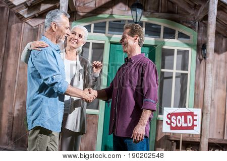Happy Senior Wife Holding Keys Of New House While Her Husband Shaking Hands With Saleman