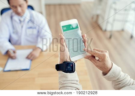 Close-up shot of wrinkled hands using activity tracker and smartphone in order to check heart rate during check-up