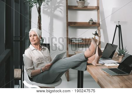 Senior Business Woman On Worklace Reading With Her Legs On Table