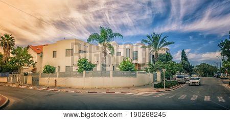 Rishon LeZion, Israel-February 18, 2016: View of modern two-story house under beautiful sky. The home is surrounded semi-circular fence, green trees and palms. Urban asphalt road in a foreground. Bright sunny day. It is located at intersection Graetz and 