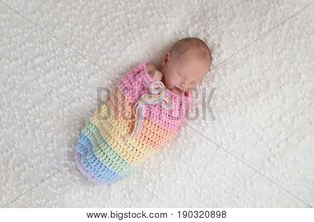 A three week old newborn baby girl bundled up in a rainbow colored snuggle sack. She is lying on a white bouncle blanket.