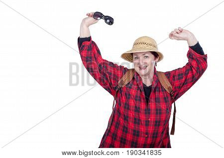 Funny older woman with backpack poses on white background. Tireless woman wears red plaid shirt and yellow straw hat. Her right hand holds sunglasses. Elderly people traveling concept