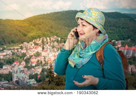Older lively woman talks by mobile phone in mountains. Backpacking woman standing above on blurred highland city background. She wears turquoise clothes and colored round hat. Older people traveling concept