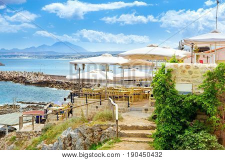 Outdoor resort cafe with white parasols on top of hill above highway along promenade Emmanouil Kefalogianni. Greek architecture on coast of Kolpos Almirou. Rethymno Greece