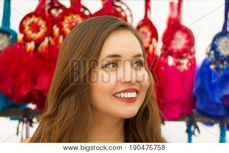 Close up of a beautiful smiling young woman looking at the horizont in front of blurred colorful catchdreamers, in colorful market background.