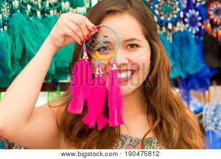 Close up of a beautiful smiling young woman pointing in from of her face a pink catchdreamers, in colorful market fabrics background.