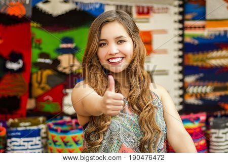 Beautiful smiling young woman with humbs up and posing for camera, with colorful andean traditional clothing fabrics background.