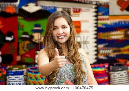 Beautiful smiling young woman with humbs up and posing for camera, with colorful andean traditional clothing fabrics background.