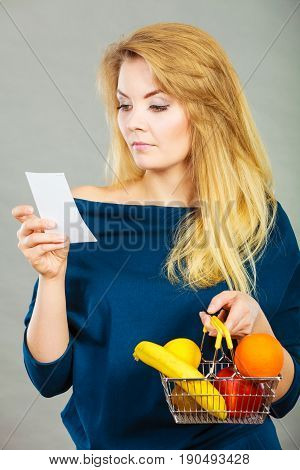 Worried woman holding shopping basket with fruits looking at bill receipt being scared of huge prices