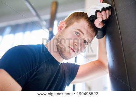 Low angle portrait of young tired sportsman leaning on punching bag during boxing practice in fight club and looking at camera