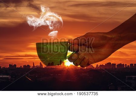 Man hand holding a cup of hot coffee aroma drifting far on the background silhouette of the city with sunriseConcept Double exposure