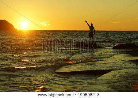 Silhouette of a man standing on a rock with his hand raised upwards with his fingers spread out against the sunset on the beach.