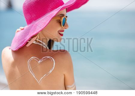 Back view on a beautiful woman in pink hat with sunscreen heart shape on her shoulder having a sunbath on the beach