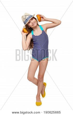 Llittle girl in swimsuit and summer hat having fun with half oranges making fake headphones, looking at camera, over white background