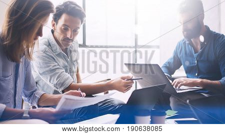 Group of three young coworkers working together at modern coworking studio.Woman pointing hand on paper document and talking with man about new startup project.Horizontal, blurred background