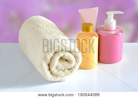 Natural hypoallergenic herbal Cosmetic products for skin care: plastic dispenser with liquid soap bottle of lotion tube of cream and white terry cotton towel on a pink floral blurred background