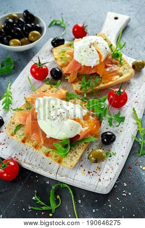Poached egg on grilled toast with smoked salmon, rucola, olives and vegetables on white board. healthy breakfast.