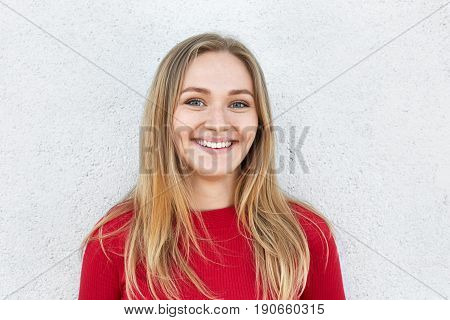 Cheerful blonde woman with freckled face charming eyes and sincere smile having pleased look while standing against white concrete wall. Bright lovely female with joyful expression. Positive emotions