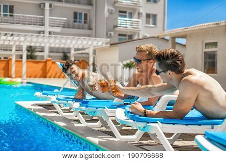 Cheers. Party at smimming pool. Young friends in swimwear and sun glasses are drinking beer while sunbathing on the chaise longue near the pool. They clink glasses