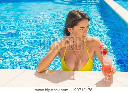 portrait of young sexy girl at swimming pool, wearing bikini. She is putting off sunglasses and looking for somebody. drinking red cocktail during vacation. Pool party