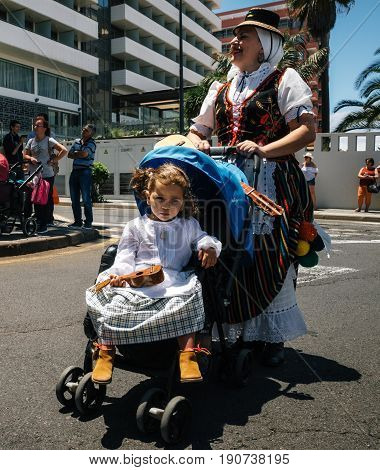 Puerto de la Cruz Tenerife Canary Islands - May 30 2017: A little girl dressed in traditional clothes sits in a stroller and holds the canary guitar Timple. Her mother in a Canarian dress. Local residents of Tenerife celebrate the Day of the Canary Island