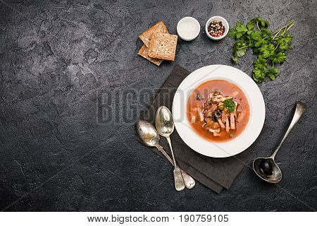 Soup with meat, olives, herbs, lemon in white bowl, black bread and spices on dark background, top view homemade food. Traditional Russian soup - solyanka