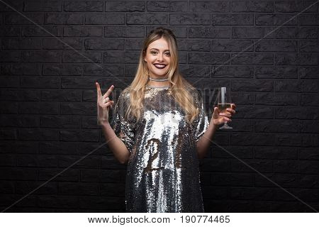 Young cheerful girl in trendy dress gesturing and holding wineglass on brick black wall.