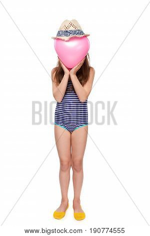 Full length child girl in swimsuit and summer hat holding pink balloon heart shape covering face, studio portrait