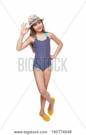 Full length child girl in swimsuit and summer hat gesturing OK sign, over white background