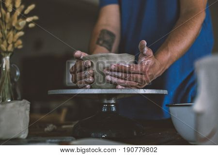 Close up of male hands working with clay on artisan working stand from dry wood surrounded by artistic workshop tools and implements creates poterry diy
