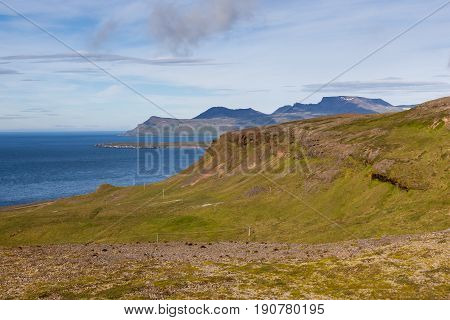 Icelandic Landscape With Green Hills On The Foreground And High Cliffs And Atlantic Ocean On The Bac
