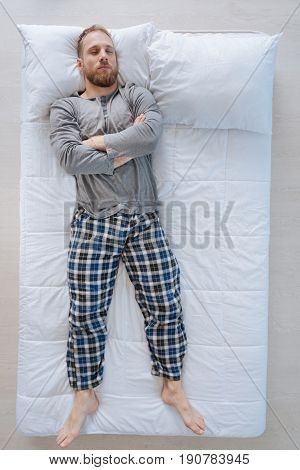 Pleasant relaxation. Serious confident adult man lying on the bed and crossing his hands while being asleep
