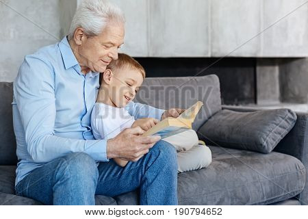 Family leisure. Nice affectionate intelligent man spending time with his grandchild and telling him fascinating stories while they cozily sitting on a couch