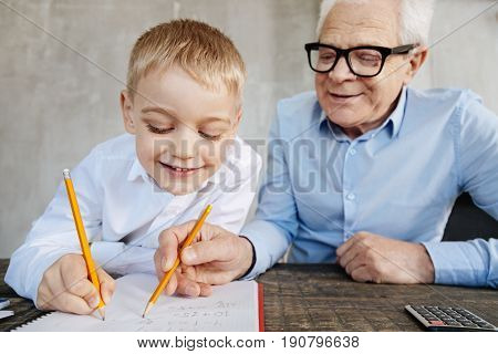 Sharing his wisdom. Emotional gifted adorable kid fulfilling his home assignment while his grandpa watching him doing it and correcting his mistakes