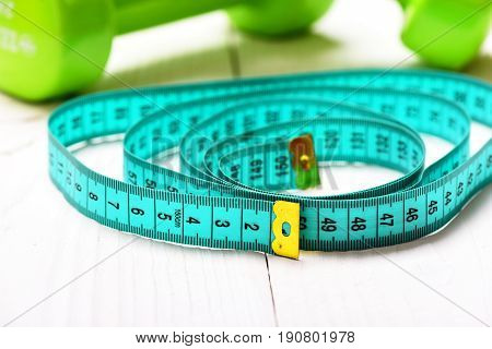 Sports fitness equipment. Cyan measuring tape with golden ends on light wooden surface and green dumbbells on background close up and selective focus. Workout and healthy lifestyle concept