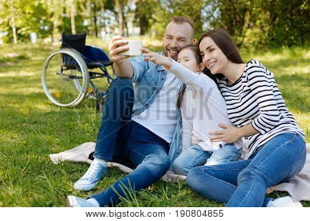 Time for photo. Smiling bearded man holding phone in right hand and sitting on the plaid with his wife and daughter while doing selfie