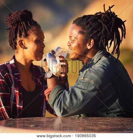 Interracial couple making a toast with interwined arms while looking into each others eyes with cheerful smiles as they enjoy their first outdoor date while seated at a public picnic table in Africa.