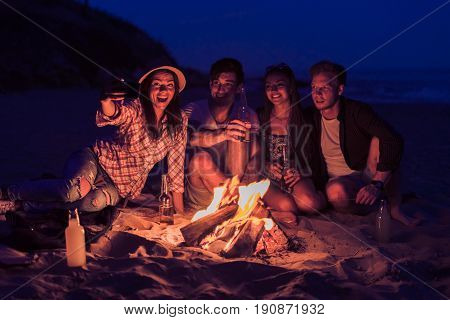 young and cheerful friends sitting on beach and take selfie near bonfire One man is drinking beer. Happy lifestyle
