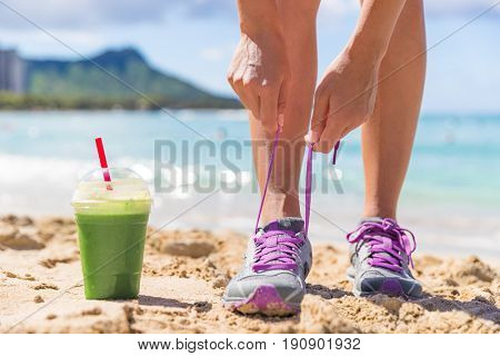 Green smoothie runner fitness woman tying running shoes at beach workout. Healthy lifestyle detox weight loss girl lacing trainers on Waikiki, Honolulu, Hawaii.