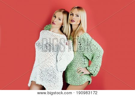 twins. Two pretty girls or cute women twins fashionable models with blond long hair stylish makeup and sexy knitted sweaters posing on red background. Female fashion and beauty