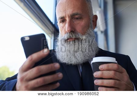 Close-up of pokerfaced man age of 50-60 holding cup of coffee and surfing in phone