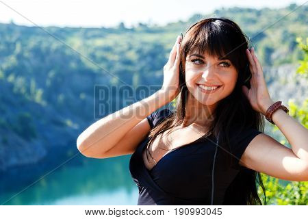 Prtty cheerful young woman listening to music in headphones outdoors. walking on a mountains at sunny summer day.