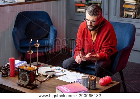 Bearded Astrologist Sitting In Armchair Reading Book And Doing Practice