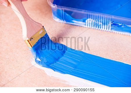 The Hand With A Brush Paints With Blue Acrylic Paint A Brown Fiberboard Surface