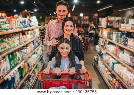 Young Parents And Daughter In Grocery Store. They Walk Among Hygeine Shelfs And Smile On Camera. Gir